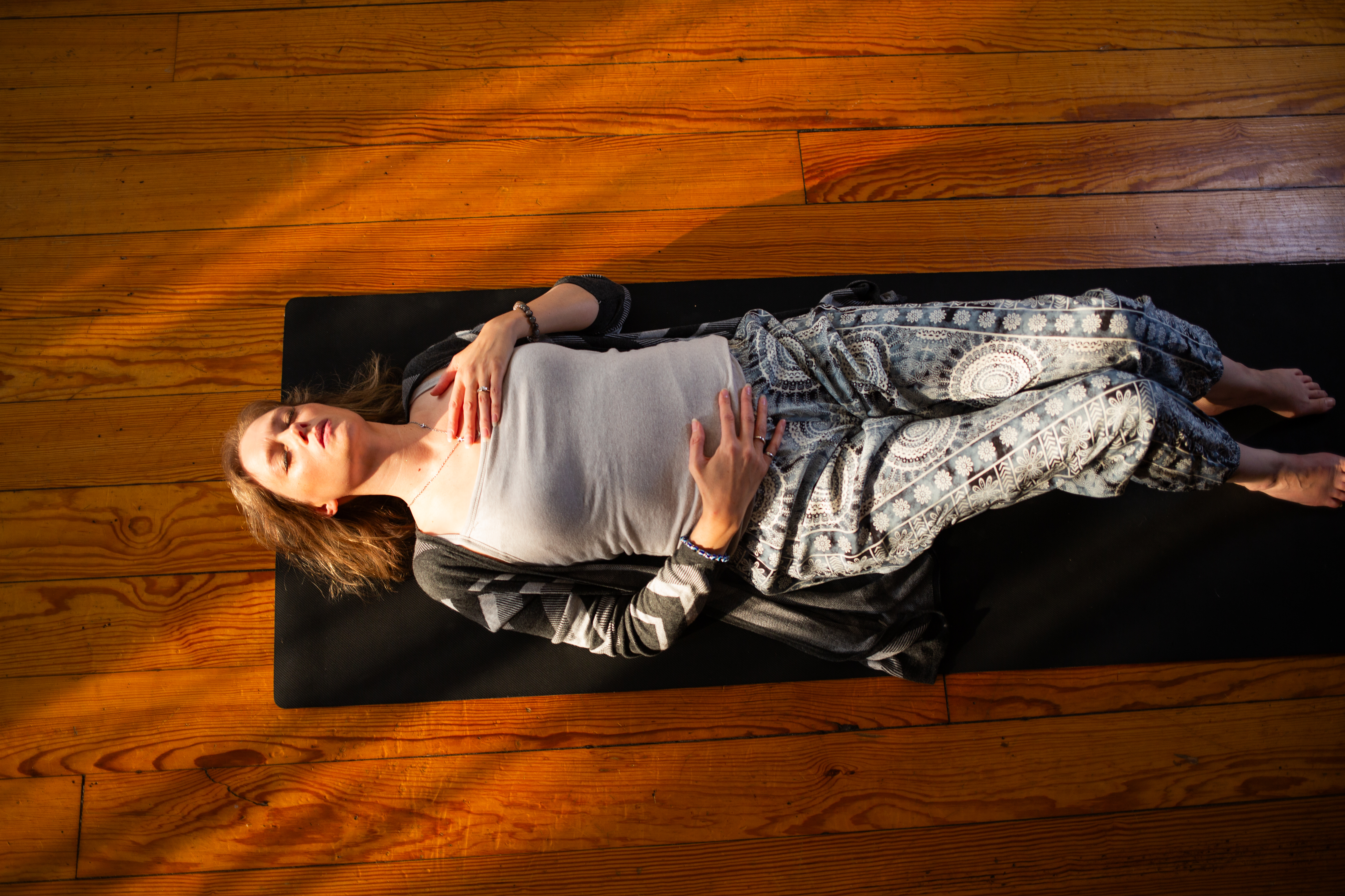 Picture of a female laying down on a mat on a wood floor, her left hand on her chest and right hand on her stomach