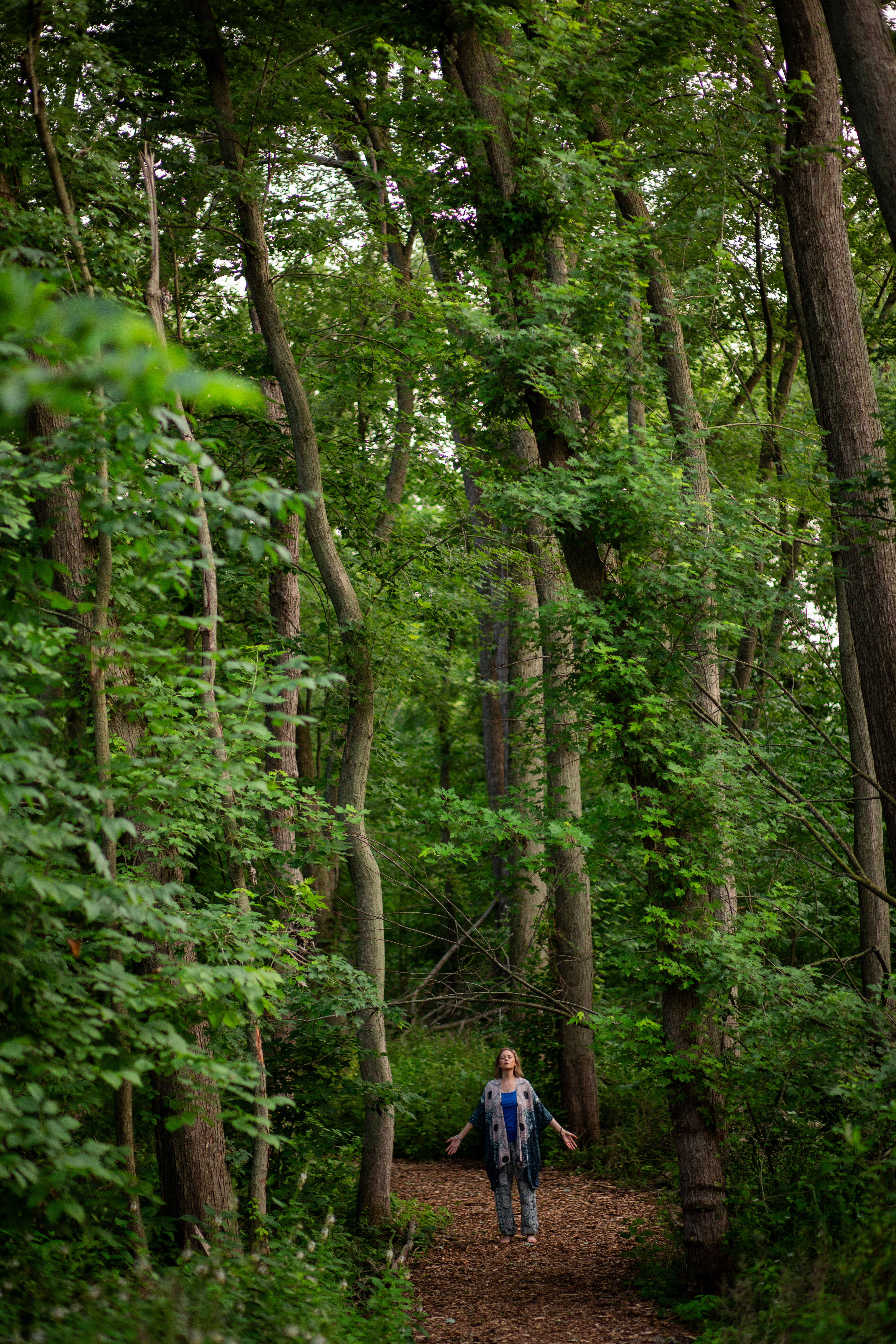 Picture of a female standing arms open wide looking up to the sky in the  middle of a path in a forest