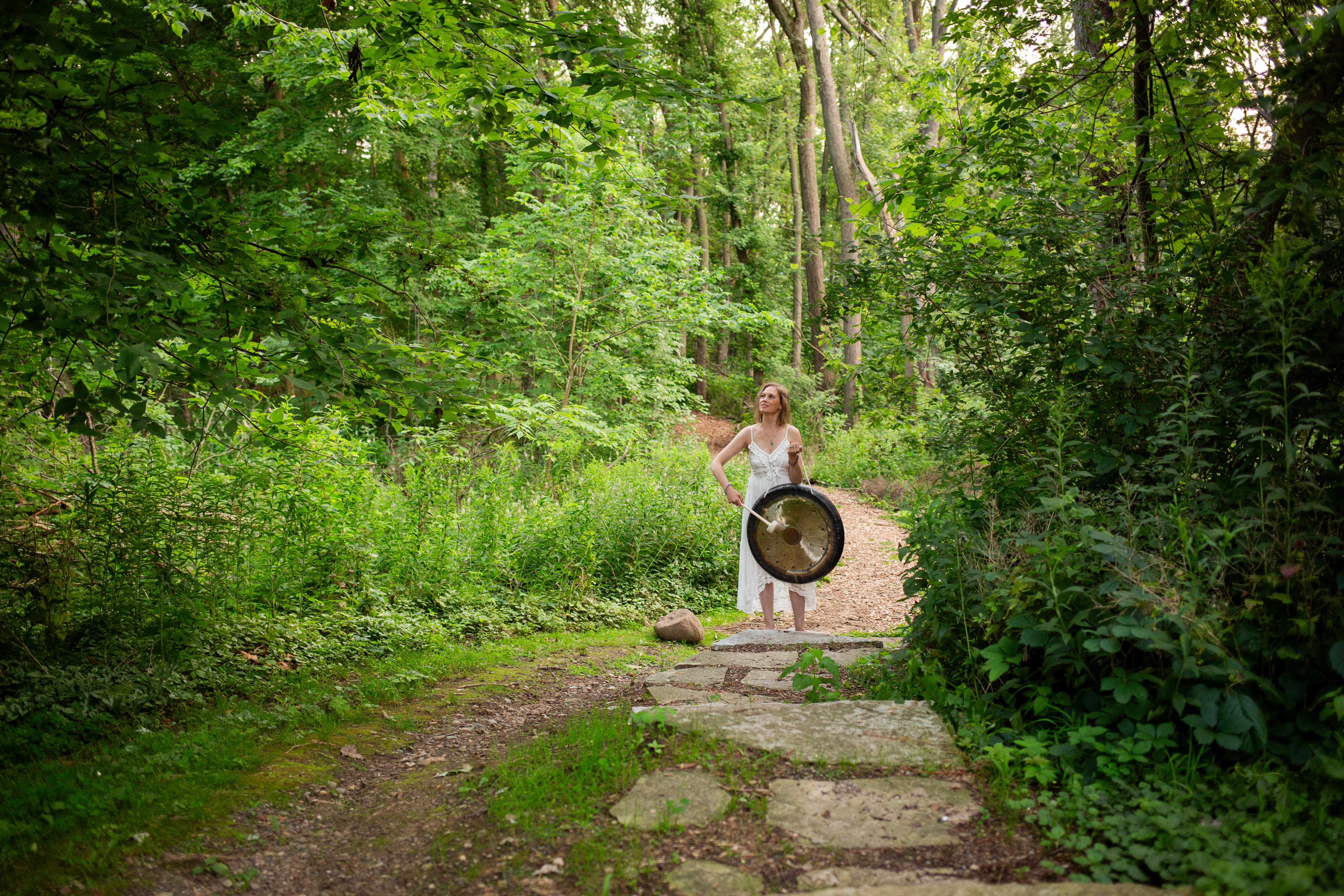 Picture of a female playing a gong in the middle of a path in a forest