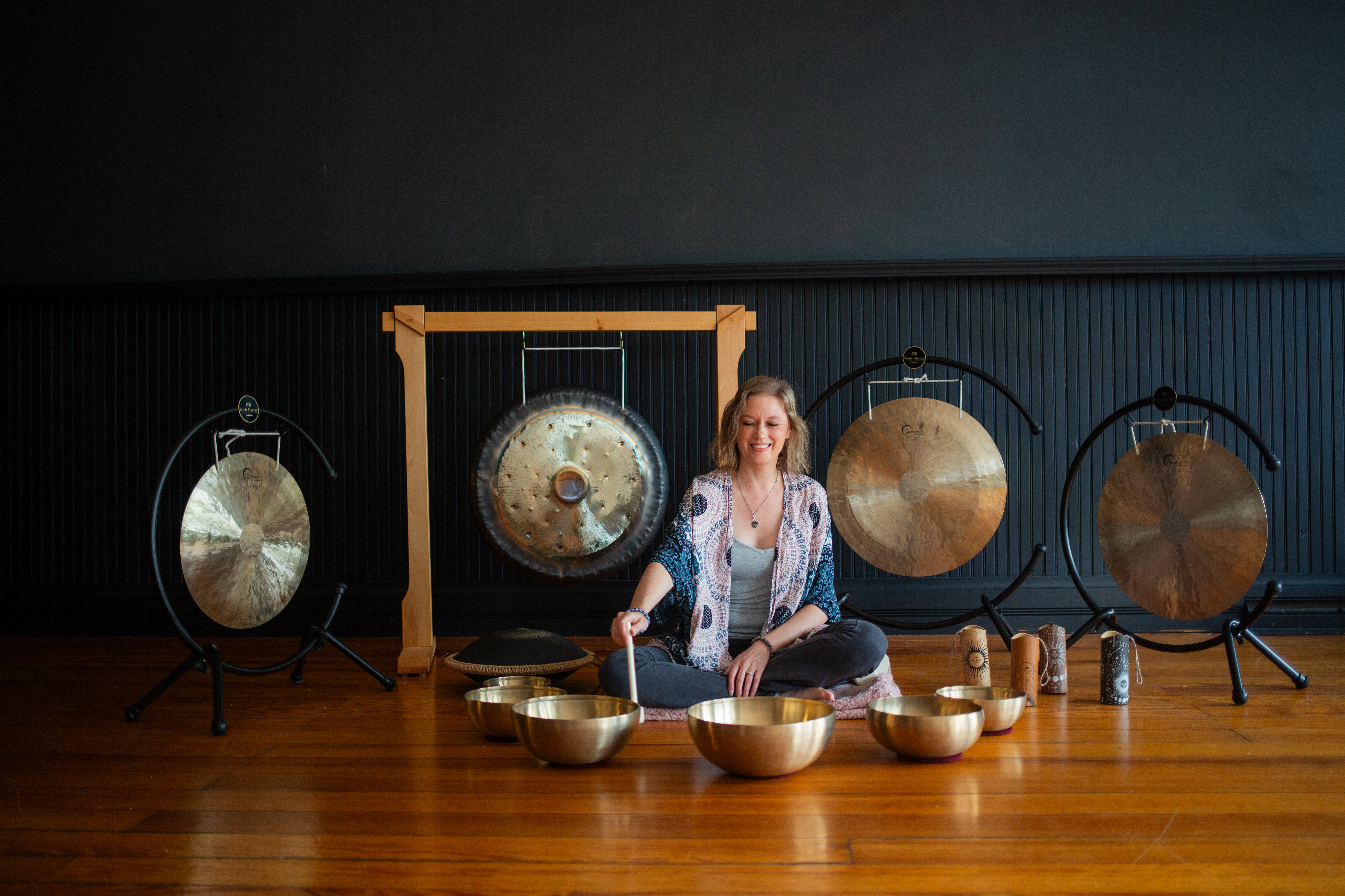 Female sitting on a pink blanket playing Tibetan bowls, with Cosmic chimes to the right, four gongs behind her and a tongue drum to the left